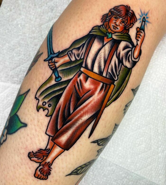 "Colorful tattoo of a hobbit from Lord of the Rings with a glowing sword and leaf cape on a person's leg."
