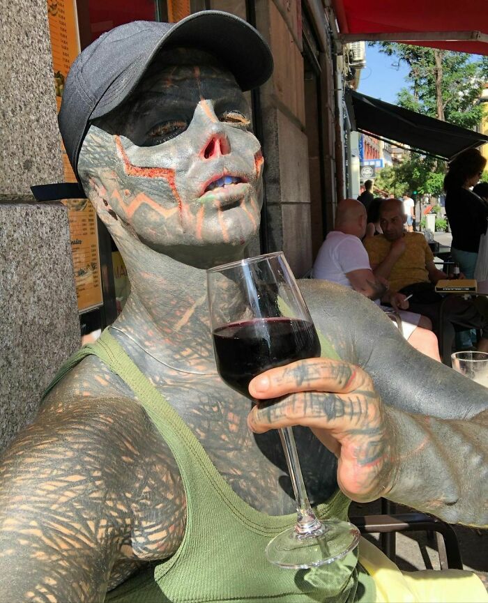 "Black Alien" with full-body tattoos holds a wine glass, enjoying a sunny day at an outdoor café. "Black Alien" with full-body tattoos holds a wine glass, enjoying a sunny day at an outdoor café.