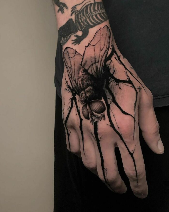 Hand with detailed gothic tattoo of a large fly and a skeletal design on the wrist.