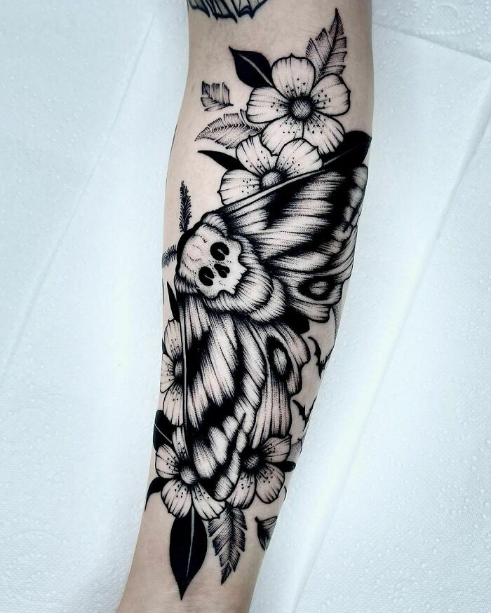 Gothic tattoo featuring a death moth and flowers on forearm, showcasing intricate black ink details.
