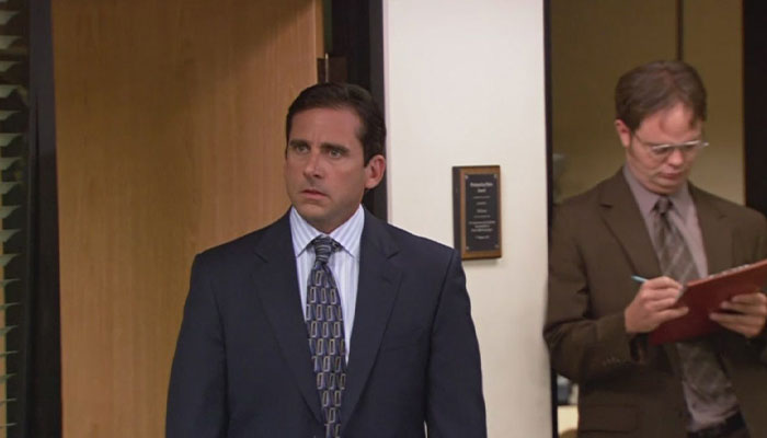 micheal scott looking lost while dwight schrute is standing in the background and writing something down