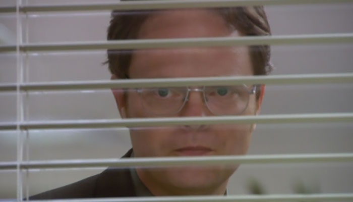 Dwight Schrute with a scary look on his face looking through blinds