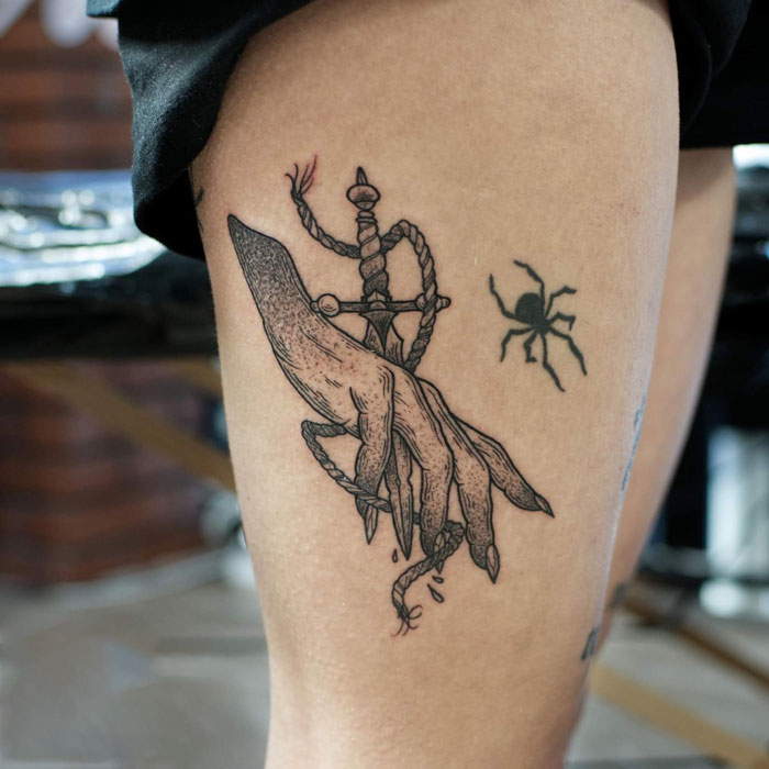 Gothic tattoo featuring a dagger entangled with a hand and a spider on a person's thigh.
