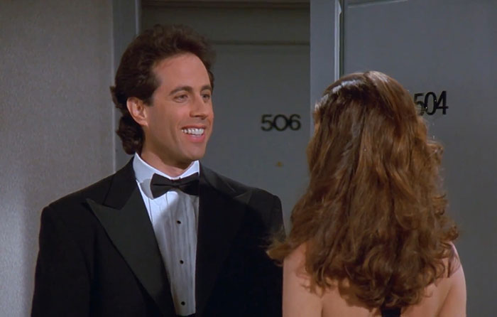 Jerry Seinfeld in a black suit smiling and talking to a woman in a scene featuring funniest Seinfeld quotes about life.