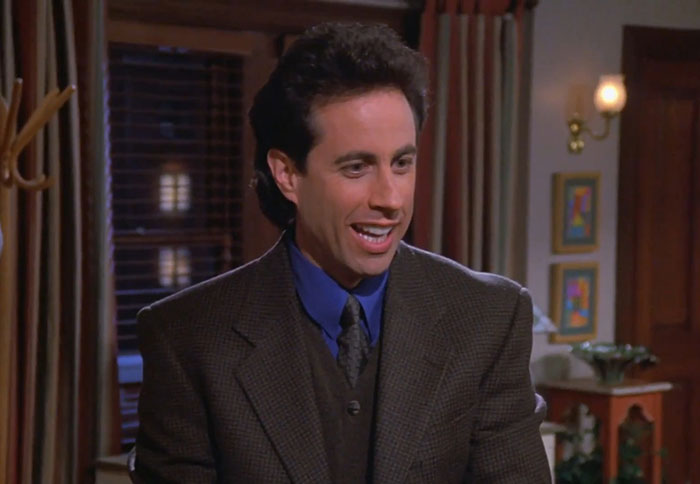 Jerry Seinfeld wearing a brown suit smiling in a living room, illustrating funniest Seinfeld quotes about everyday life.