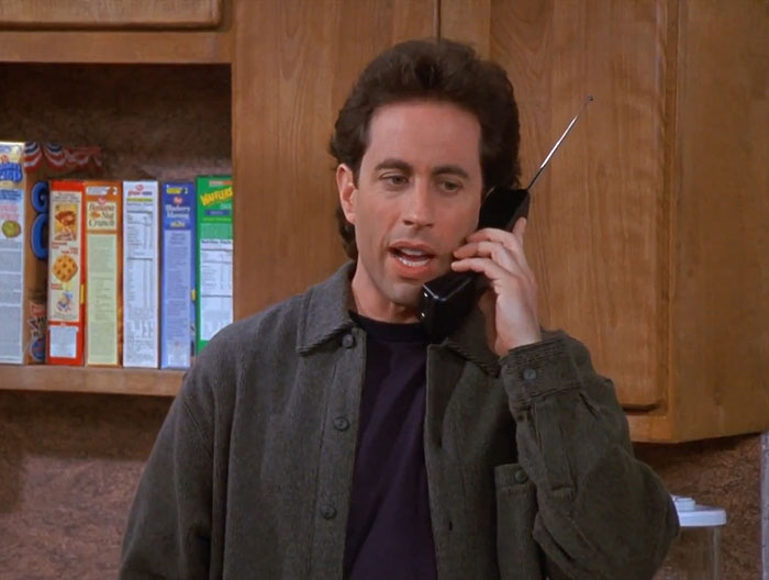 Jerry wearing a gray sweater and dark blue shirt, holding a phone, illustrating funniest Seinfeld quotes about everyday life.