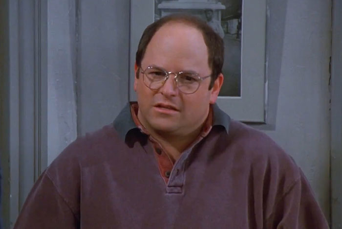 George wearing a purple sweater from Seinfeld, one of the funniest Seinfeld quotes moments that sum up everyday life.