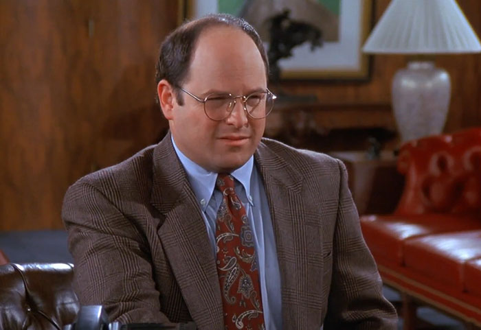 George wearing a brown suit and blue shirt, featured in funniest Seinfeld quotes that sum up everyday life.