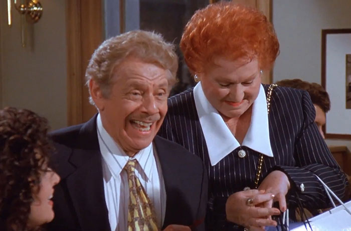 Frank Costanza in a black suit laughing with a woman, capturing a memorable Seinfeld quote about everyday life.