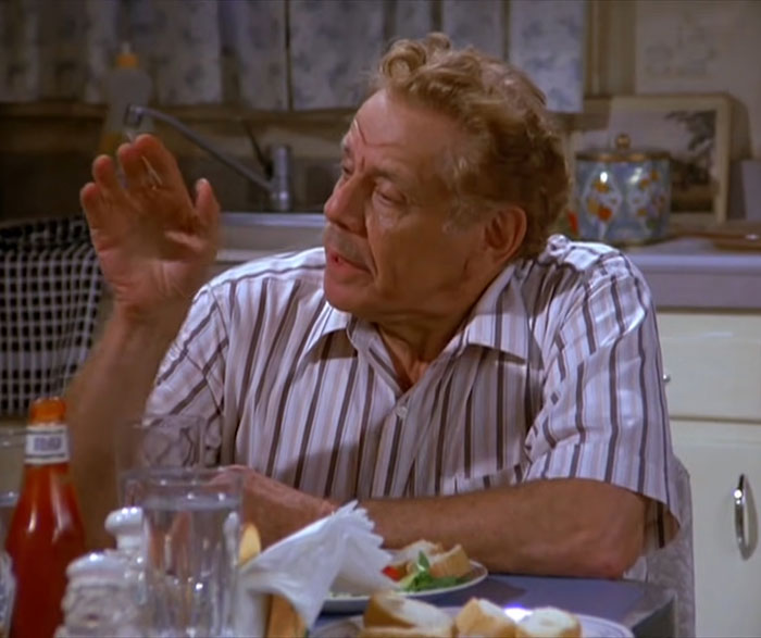 Frank Costanza in a white shirt at a kitchen table, expressing a classic Seinfeld quote about everyday life.