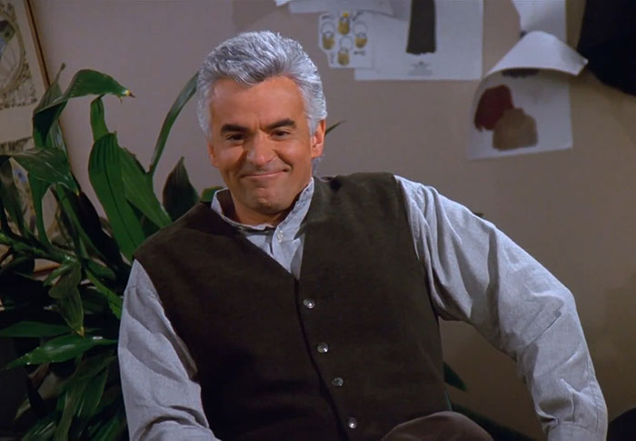 J. Peterman wearing brown vest and blue shirt, smiling in a casual setting, capturing funniest Seinfeld quotes vibe.