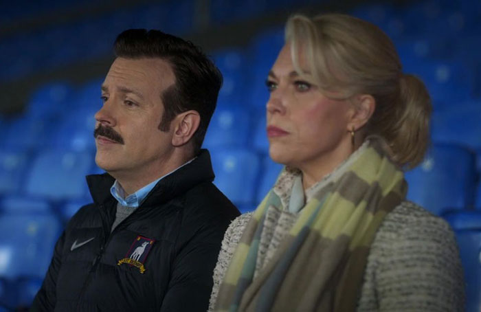 Ted Lasso wearing jacket and standing with woman