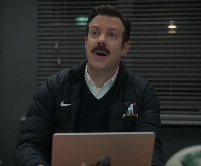 Ted Lasso wearing blue sweater and white shirt, jacket