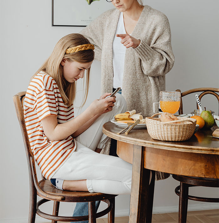 Mom looks upset while her 14-year-old daughter ignores her, focused on a phone at the kitchen table during breakfast. Mom looks upset while her 14-year-old daughter ignores her, focused on a phone at the kitchen table during breakfast.