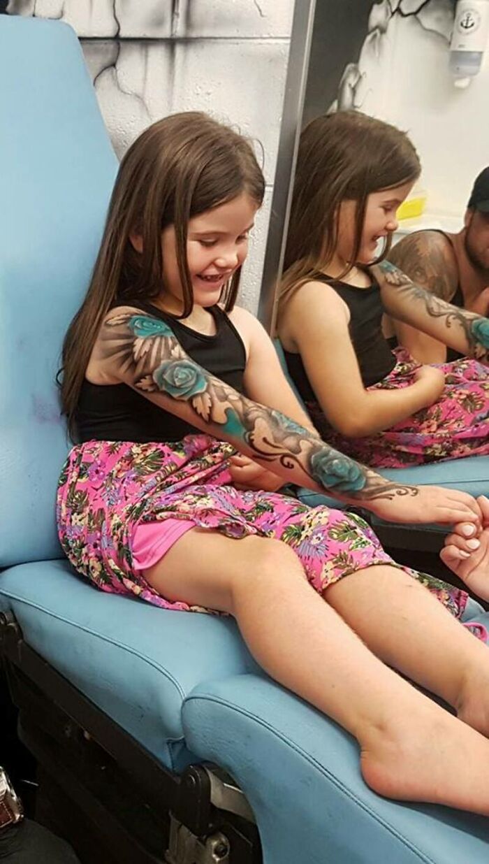 A child smiles while admiring a spray-on tattoo on her arm, sitting in a hospital chair.