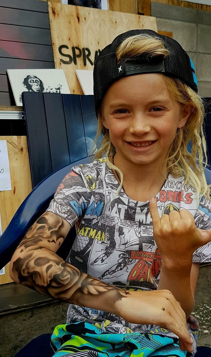 Child showing spray-on tattoos with a big smile, wearing a comic-themed shirt and cap.