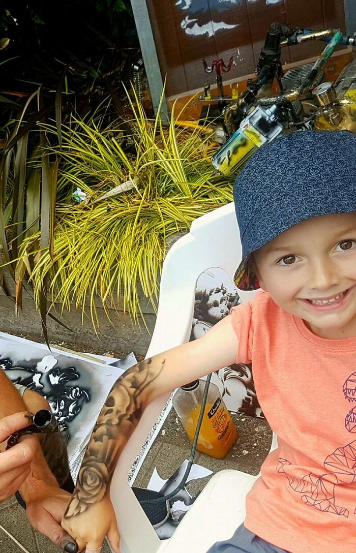 Child with spray-on tattoos, wearing a hat and smiling, receiving body art from an artist to brighten their day.