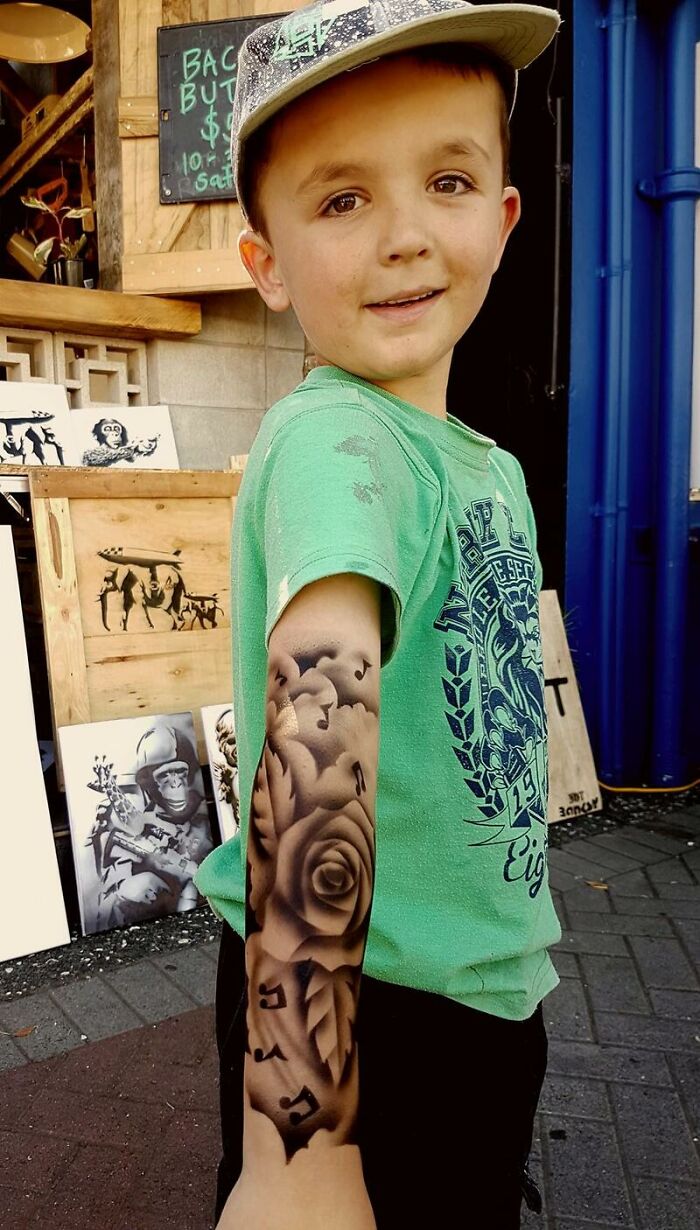 Child with spray-on tattoo featuring a rose and musical notes on the arm, wearing a green shirt and hat.