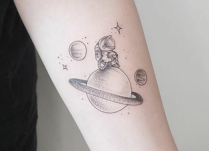 Tiny astronaut sitting on saturn arm tattoo