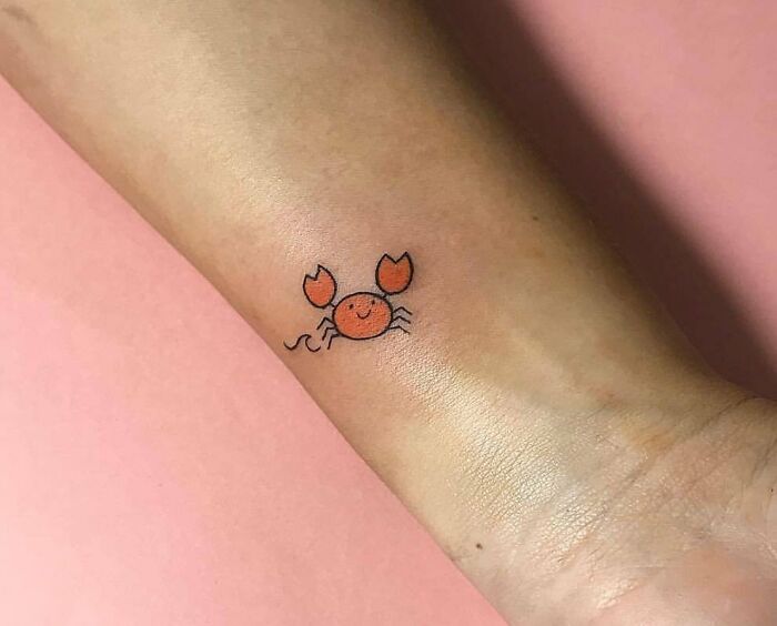 Cute crab zodiac tattoo on arm, a minimalistic design with orange color, set against a pink background.