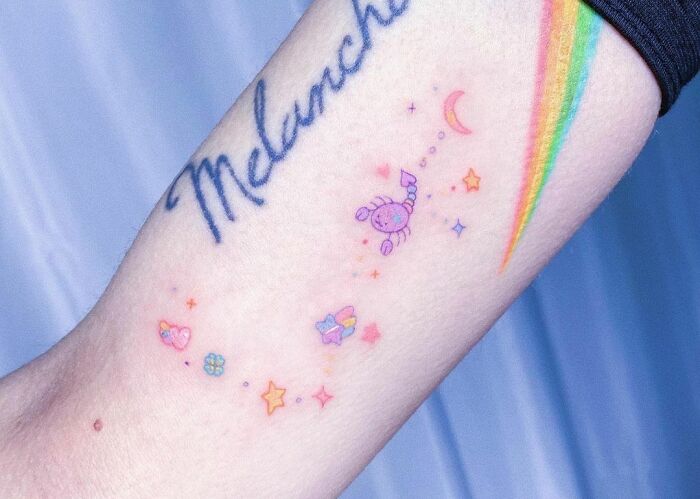 Colorful zodiac tattoo featuring stars, hearts, and a scorpion on an arm, with rainbow and script elements.