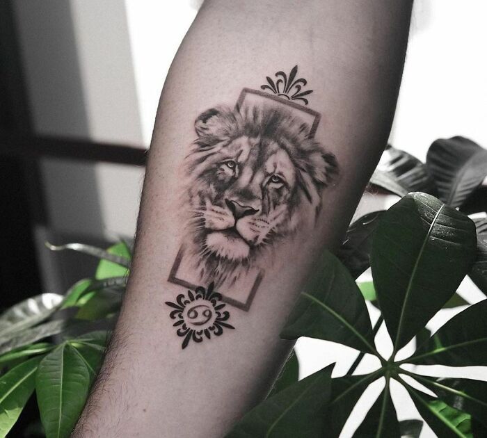 Lion zodiac tattoo on forearm, surrounded by decorative symbols with plants in the background.