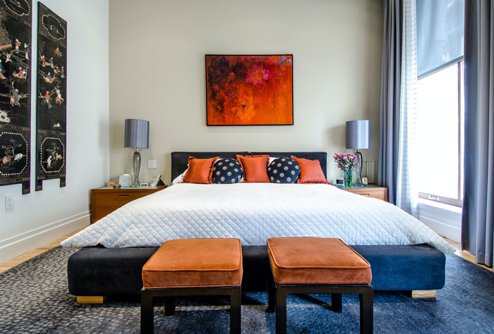 Color contrasting bedroom with orange cushions, picture on the wall, seating, and gray bed, lamp and curtains