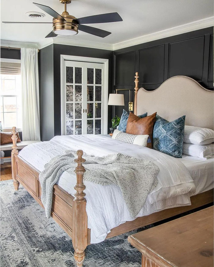 A bedroom with black walls, wooden bed platform, cushions, white bedding and black vintage ceiling fan