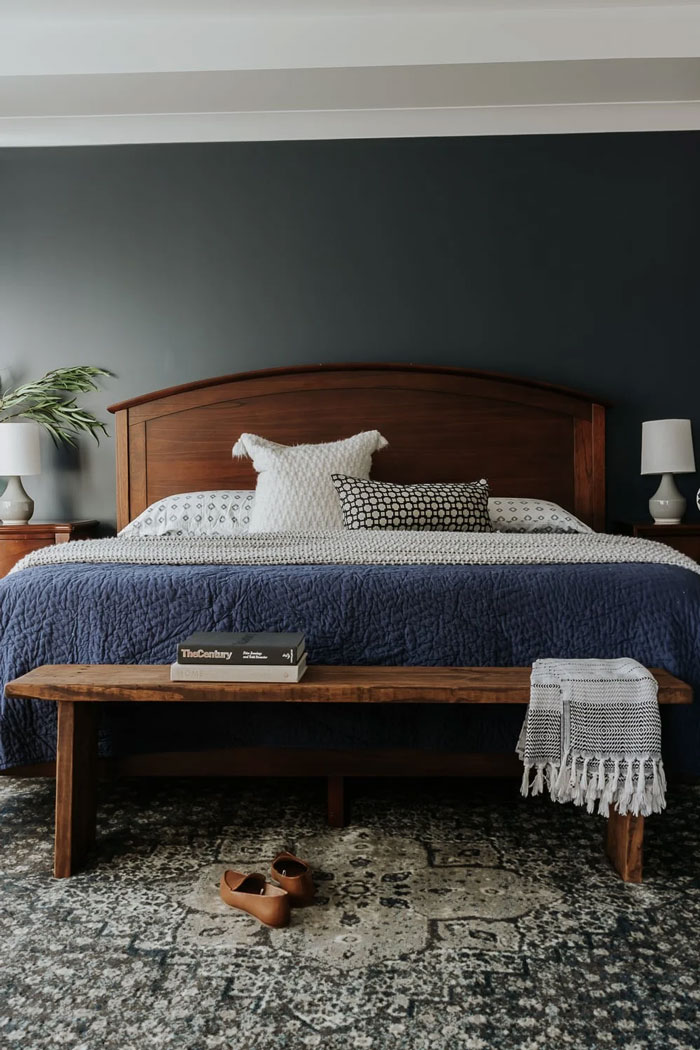 Dark colored bedroom, with blue bedding and walnut wood bench at the foot of the bed