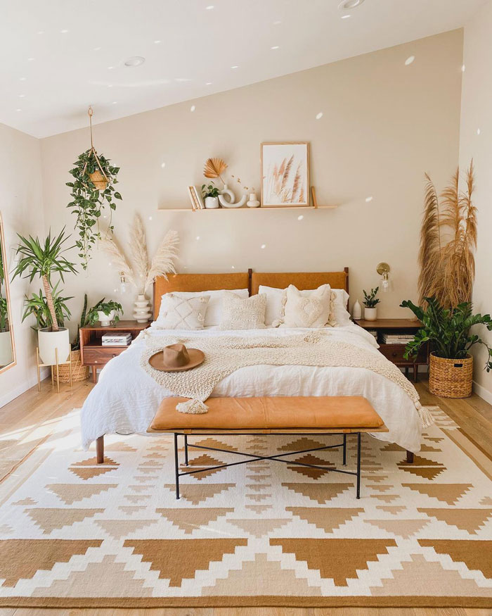 Boho style bedroom with soft beige colored walls, orange bench at the foot of the bed, white bedding and a lot of plants all over the room