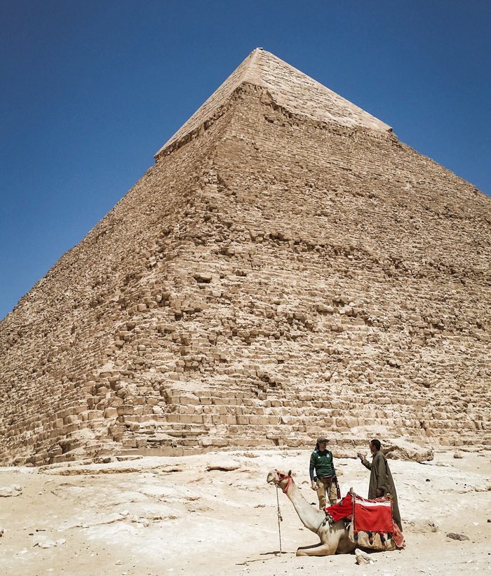 Two people and a camel in front of a pyramid