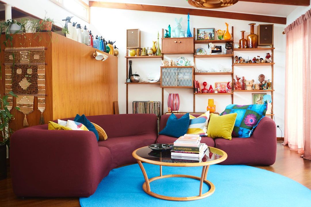 Bright living room with a large couch, colorful pillows, and a wooden shelf showcasing decorative items and books. Bright living room with a large couch, colorful pillows, and a wooden shelf showcasing decorative items and books.