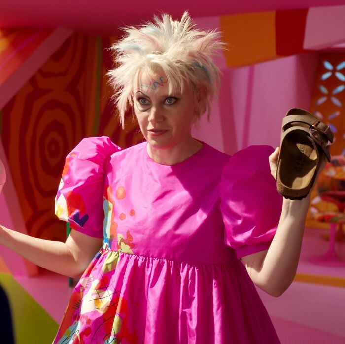 Person in a colorful outfit holding a shoe, resembling a movie-inspired ‘Weird Barbie’ character. Person in a colorful outfit holding a shoe, resembling a movie-inspired ‘Weird Barbie’ character.