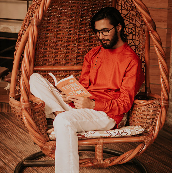 A man in an orange shirt with white pants reading a book while sitting on an oval single-seater porch swing