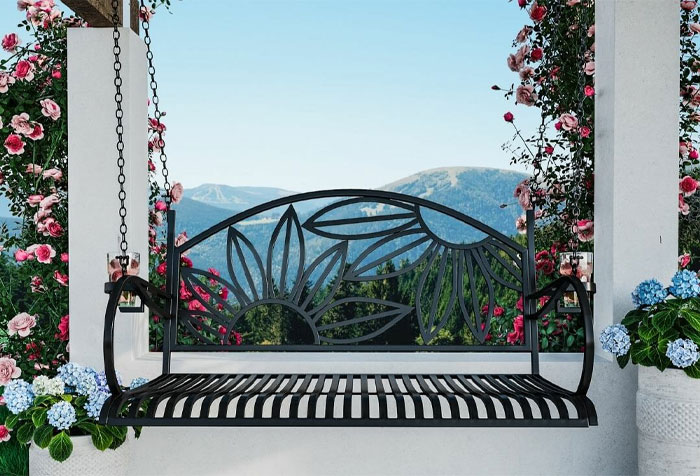 A chain-based metal porch swing with ornaments, surrounded by flowers