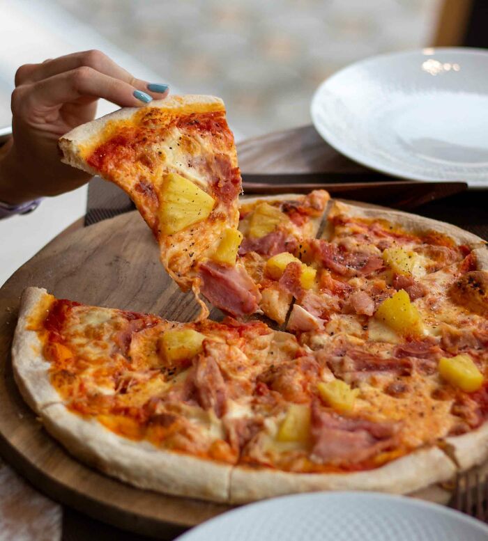A person holding a slice of Hawaiian pizza on a wooden board