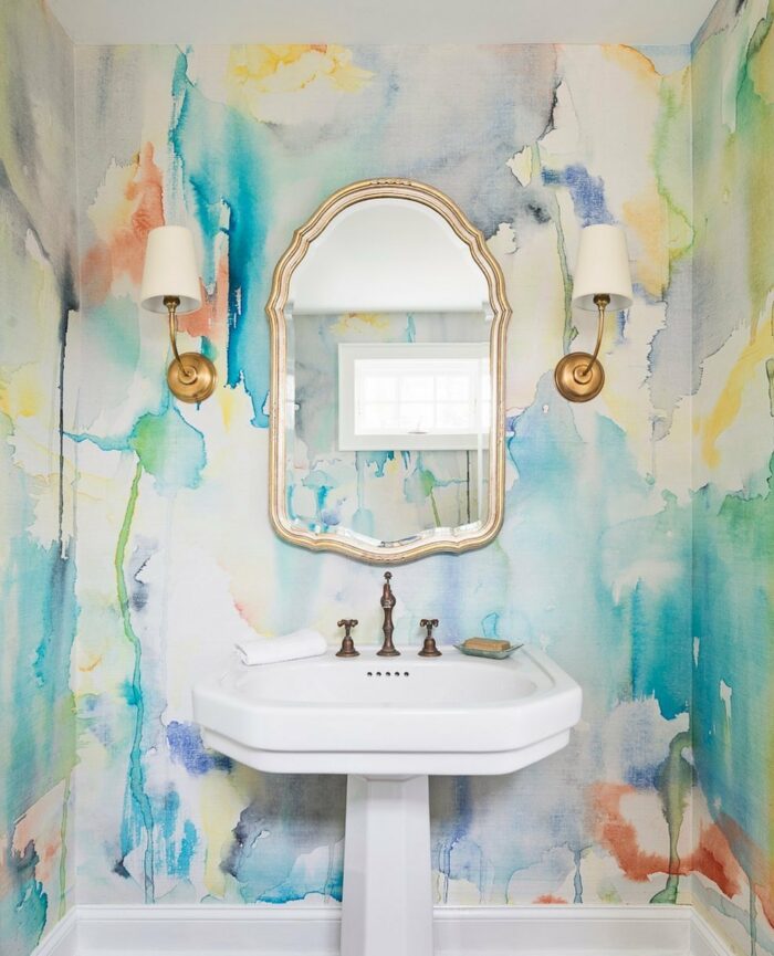 Bathroom with trendy watercolor wallpaper, white pedestal sink, ornate mirror, and gold wall sconces in a stylish space. Bathroom with trendy watercolor wallpaper, white pedestal sink, ornate mirror, and gold wall sconces in a stylish space.
