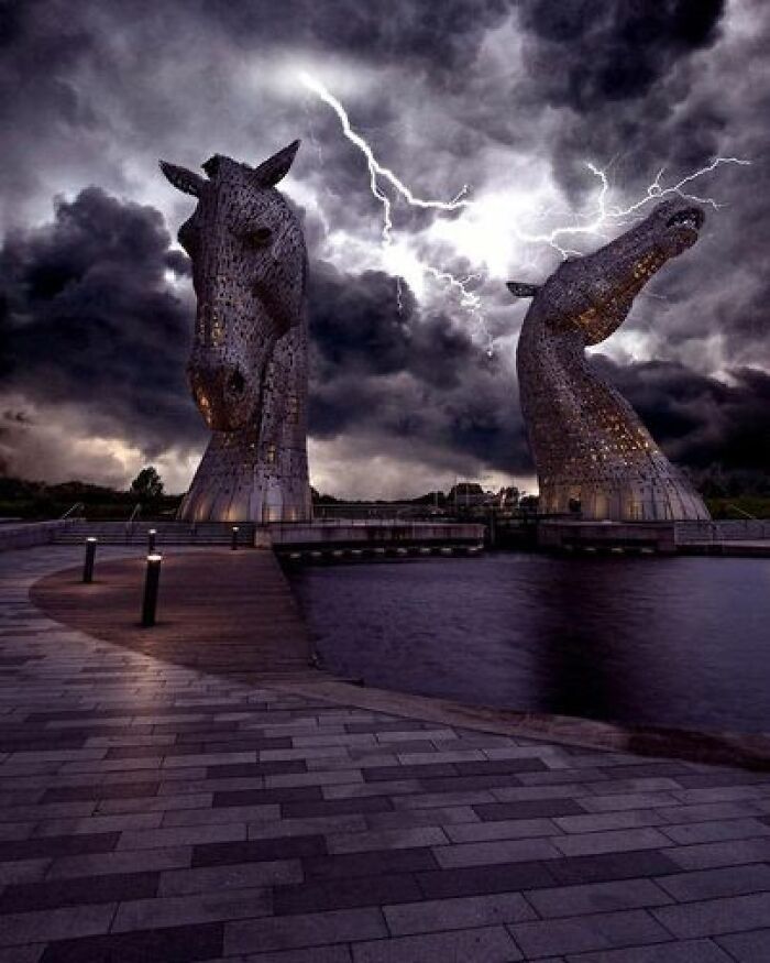 The Kelpies, Scotland, During A Thunderstorm. Rising Magnificently To A Height Of 30 Meters (98 Ft), The Awe-Inspiring Sculptures Known As The Kelpies Grace The Landscape Between Falkirk And Grangemouth. These Remarkable Horse-Head Sculptures Portray The Mythical Kelpies, Legendary Water Spirits Capable Of Shape-Shifting. Positioned Alongside The Forth And Clyde Canal's New Extension, Near The River Carron, The Kelpies Proudly Stand Within The Helix, A Visionary Parkland Project That Links Sixteen Communities In The Falkirk Council Area Of Scotland. Renowned Sculptor Andy Scott Envisioned And Brought These Sculptures To Life, With Their Completion Marking A Milestone In October 2013. Serving As A Grand Gateway At The Eastern Entrance To The Forth And Clyde Canal, The Kelpies Symbolize The Union Of The Canal And The Transformative Land Project Known As The Helix. Through Their Captivating Presence, These Sculptures Pay Homage To Scotland's Rich Horse-Powered Heritage, Leaving An Indelible Mark On The Cultural Landscape