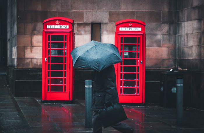 Person under umbrella walking beside red telephone booths in Manchester, United Kingdom