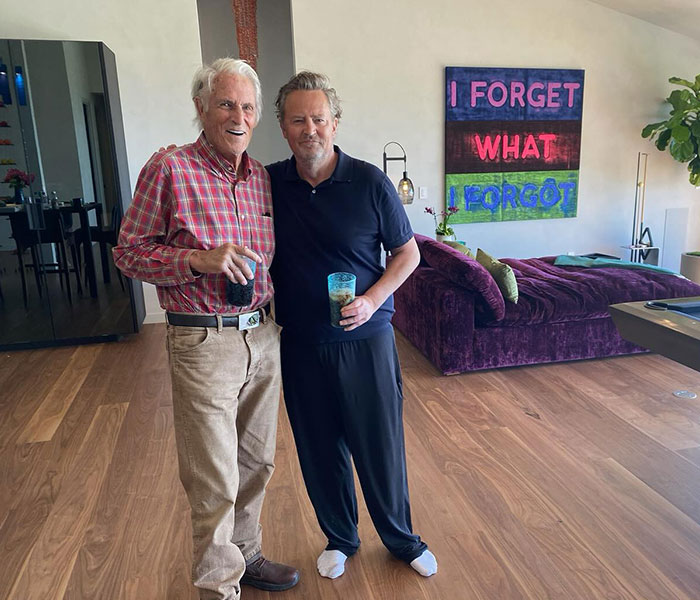 Two men standing in a living room, holding drinks, with colorful artwork in the background. Two men standing in a living room, holding drinks, with colorful artwork in the background.