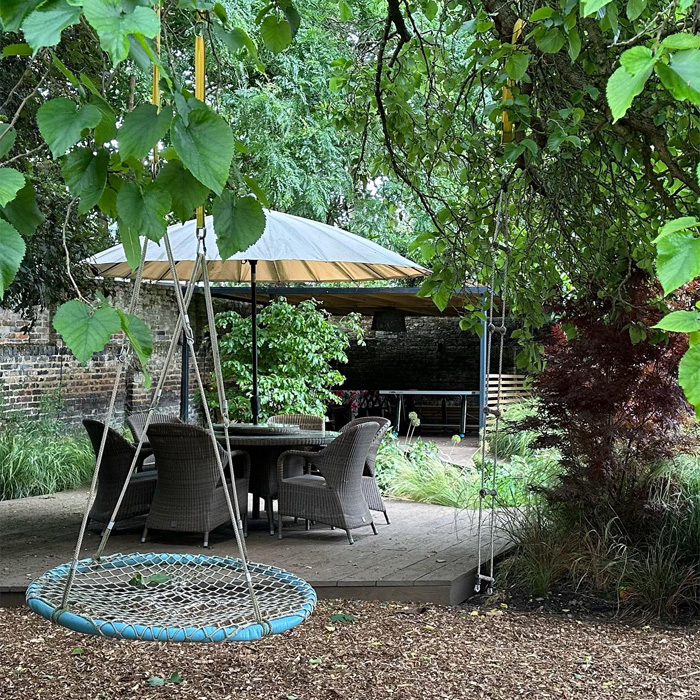 Backyard deck with wicker chairs, round table under a large umbrella, surrounded by lush trees and garden greenery. Backyard deck with wicker chairs, round table under a large umbrella, surrounded by lush trees and garden greenery.