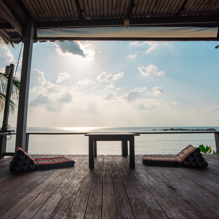 Dark brown wooden deck with floor cushions overlooking calm ocean under a bright sky, showcasing backyard deck ideas. Dark brown wooden deck with floor cushions overlooking calm ocean under a bright sky, showcasing backyard deck ideas.