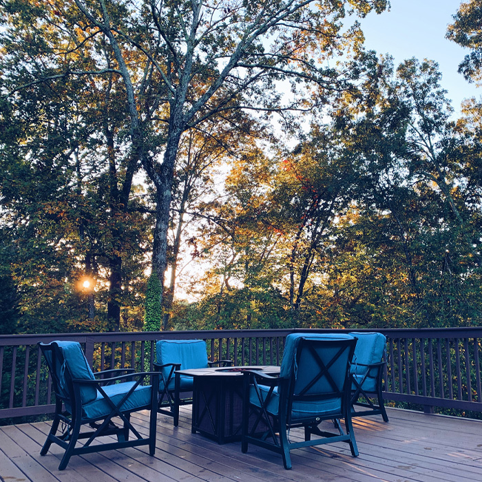 Fire pit on a backyard deck surrounded by blue cushioned chairs during sunrise with trees in the background. Fire pit on a backyard deck surrounded by blue cushioned chairs during sunrise with trees in the background.