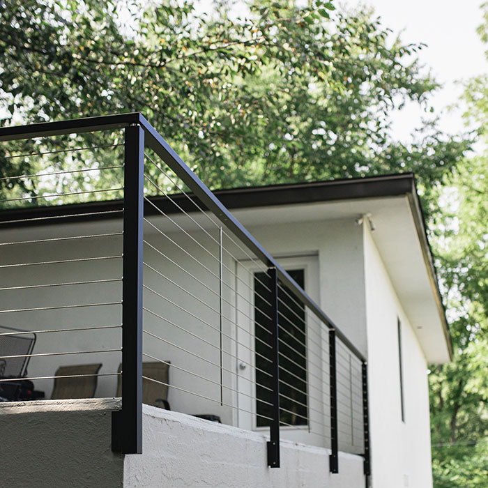 Black deck railing with horizontal cables on a white house exterior surrounded by green trees in daylight Black deck railing with horizontal cables on a white house exterior surrounded by green trees in daylight