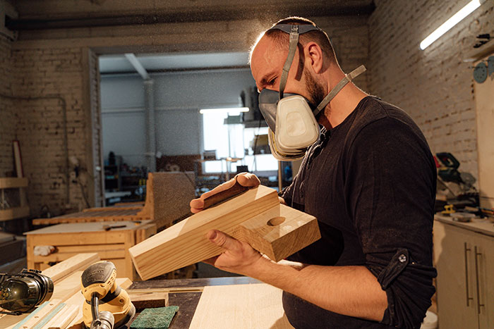 Man wearing a face mask shaping wood pieces in workshop for deck railing ideas and designs to spice up exterior spaces Man wearing a face mask shaping wood pieces in workshop for deck railing ideas and designs to spice up exterior spaces