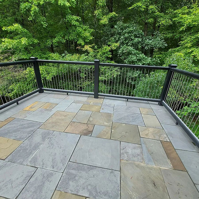 Black vertical metal deck railing with cable design on a stone porch surrounded by lush green trees. Black vertical metal deck railing with cable design on a stone porch surrounded by lush green trees.