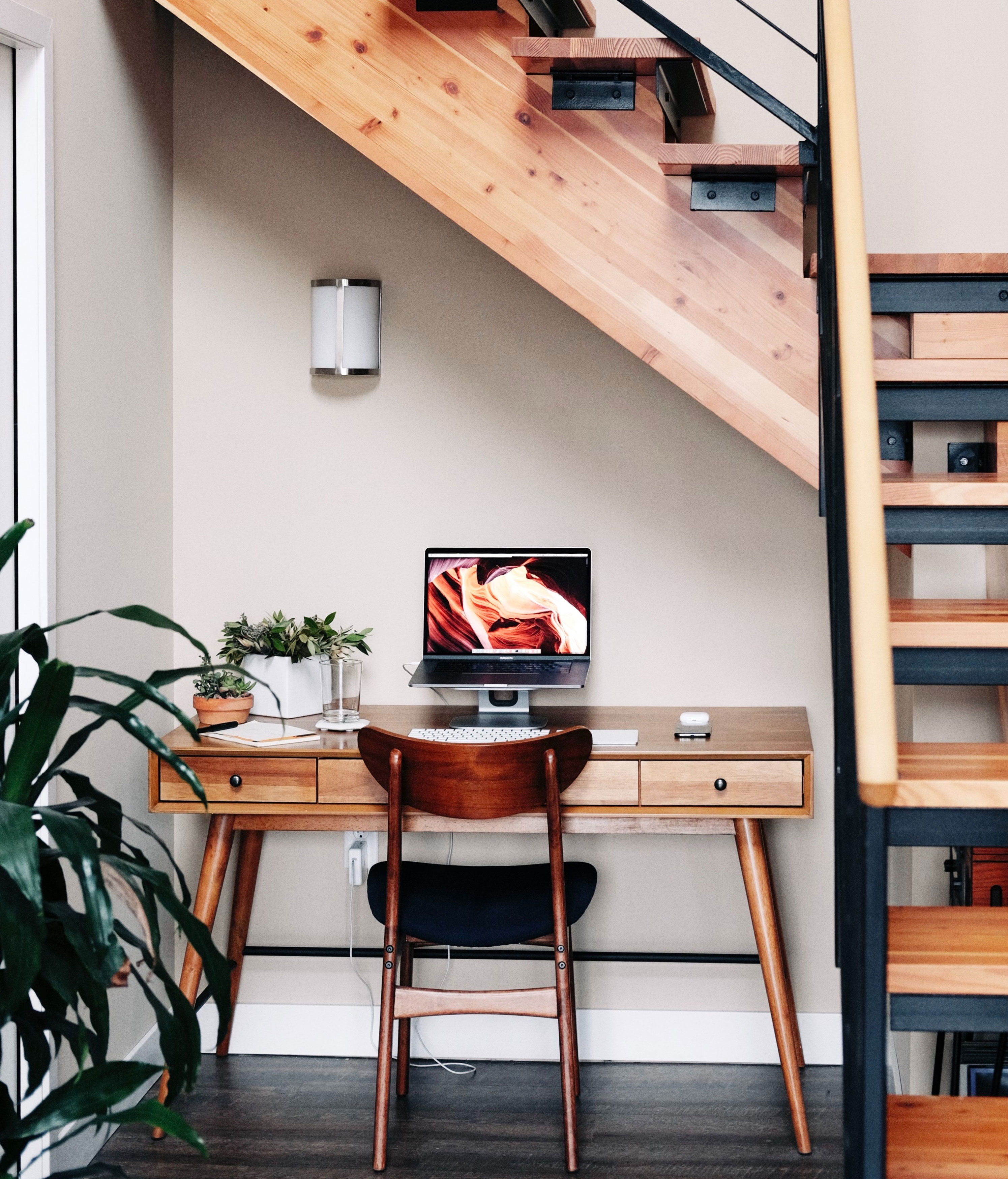 Minimalist home office idea with a laptop on a wooden desk under stairs, featuring plants for a productive workspace. Minimalist home office idea with a laptop on a wooden desk under stairs, featuring plants for a productive workspace.
