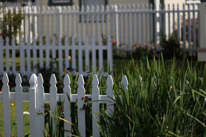 A white picket fence in a garden with grass growing near it