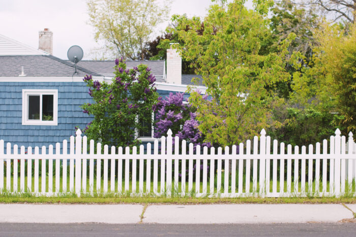 A white picket fence with a wave formation in front of a blue house and trees
