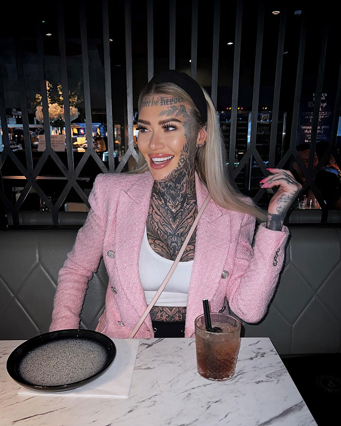 Influencer with fake forehead tattoo smiling in a pink jacket at a restaurant table. Influencer with fake forehead tattoo smiling in a pink jacket at a restaurant table.